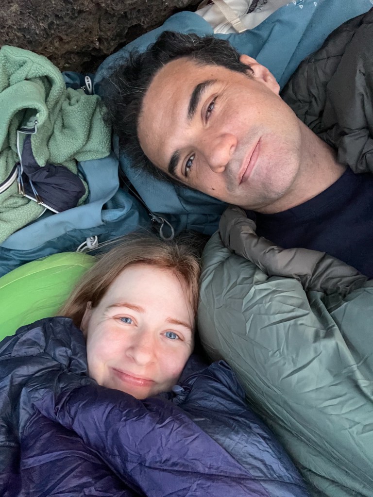 A man and a woman smiling while lying together in sleeping bags surrounded by camping gear.