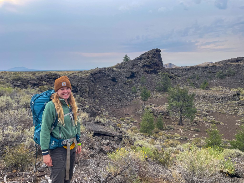 A smiling young woman wearing a brown beanie and a green jacket stands in the foreground with a large backpack. She is near a rocky area with sparse vegetation and distant hills in the background under a cloudy sky.