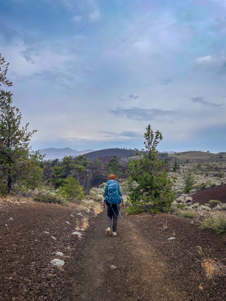 A hiker with a blue backpack walking on a trail through a volcanic landscape with trees and mountains in the background under a cloudy sky.