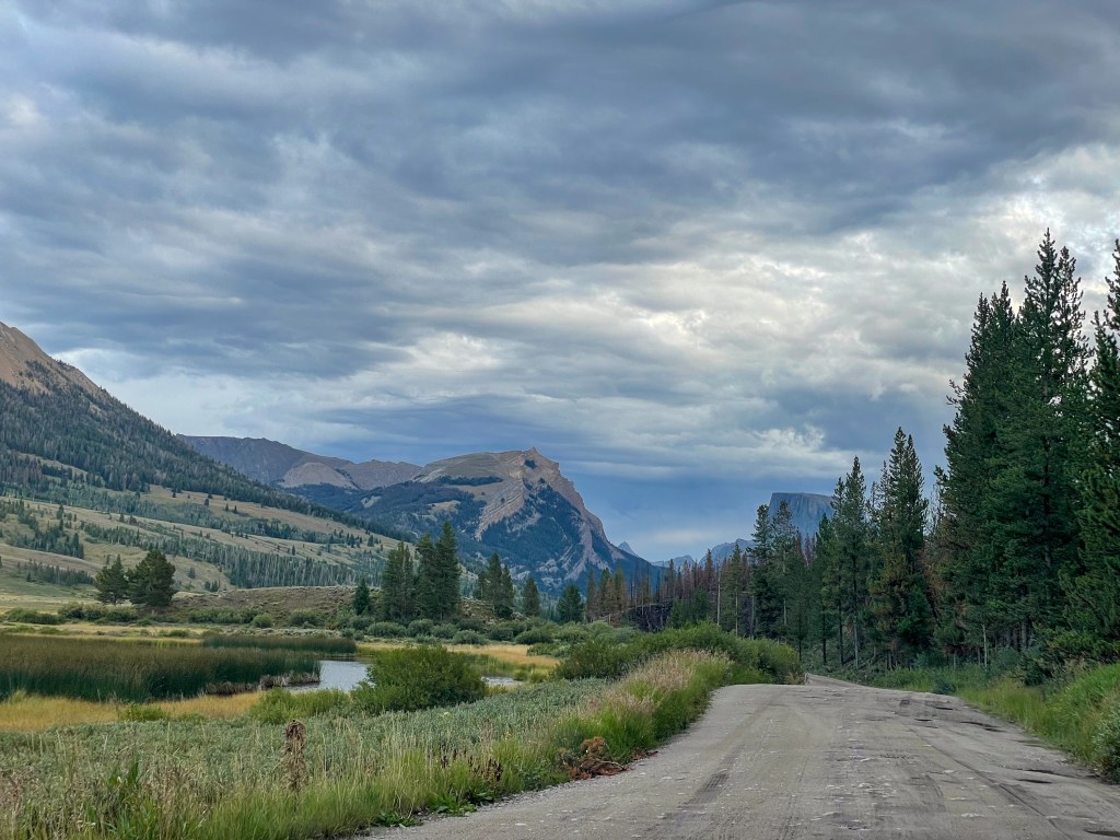 A scenic landscape featuring a dirt road surrounded by lush greenery, mountains in the background, and a cloudy sky.