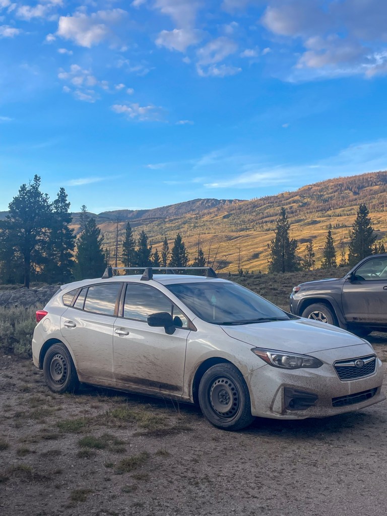 A white Subaru hatchback parked on a gravel area, showing signs of dirt and mud with mountains and a blue sky in the background.