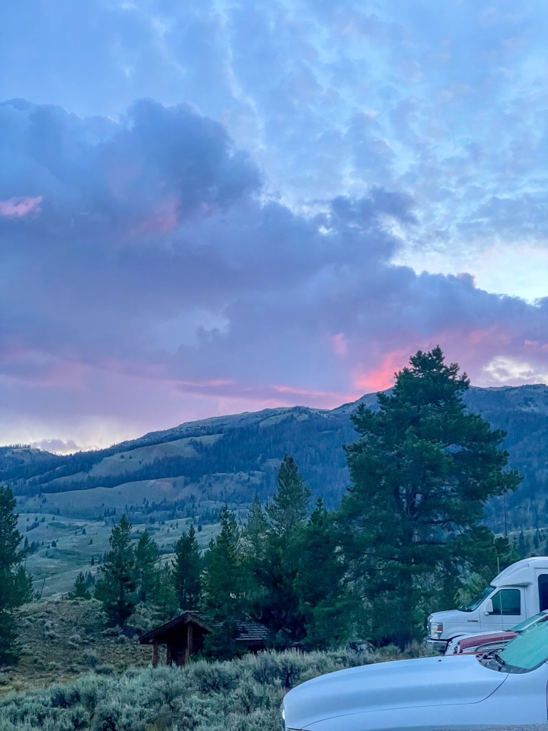 A scenic view of mountains during twilight, featuring soft pink and blue hues in the sky, with trees in the foreground and parked vehicles.