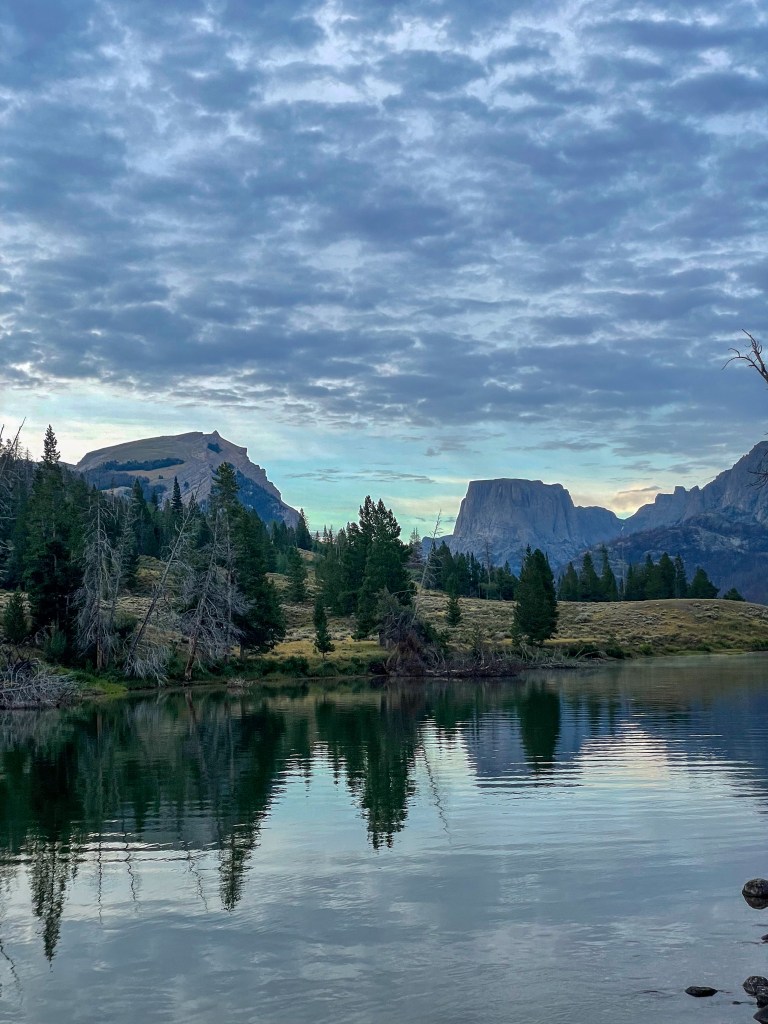 A tranquil lake reflecting mountains under a cloudy sky, surrounded by trees and grassy terrain.