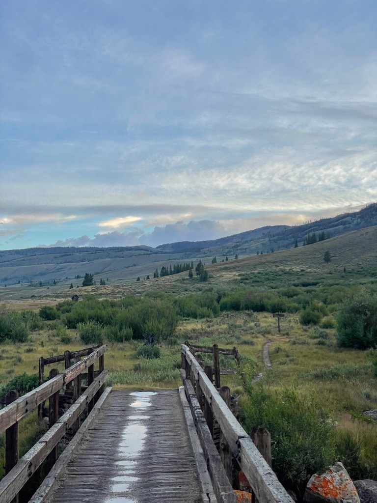 A rustic wooden bridge leading into a lush green landscape under a partly cloudy sky, with rolling hills in the distance.