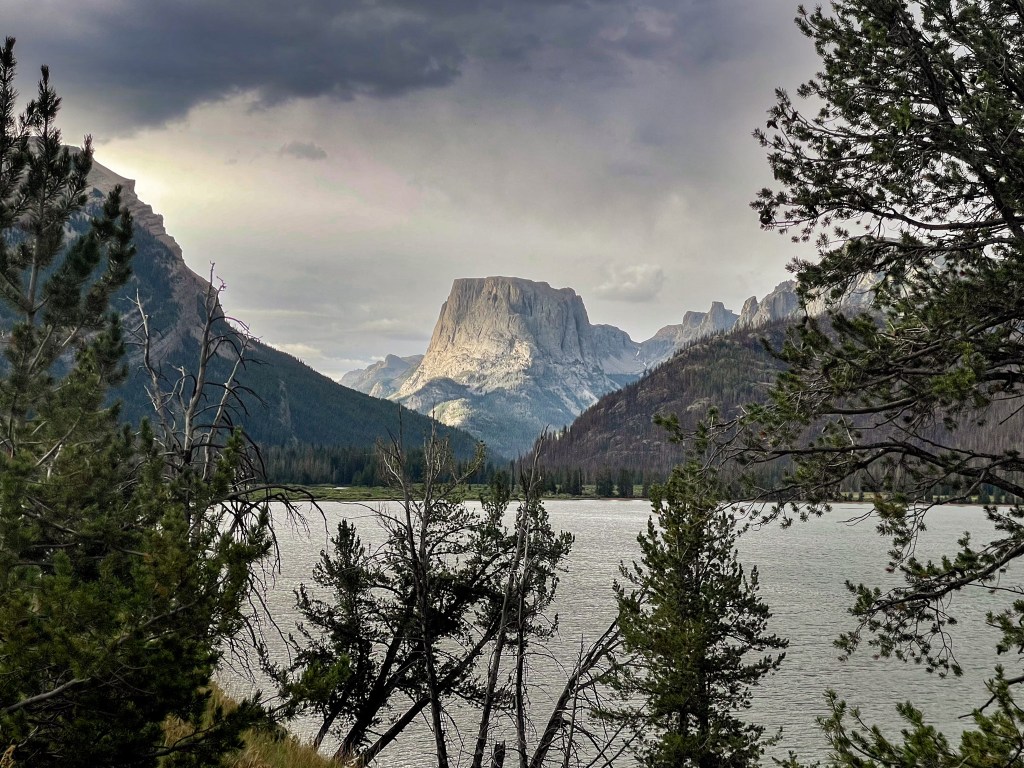 Scenic view of a lake surrounded by trees, with a prominent mountain peak in the background under a cloudy sky.