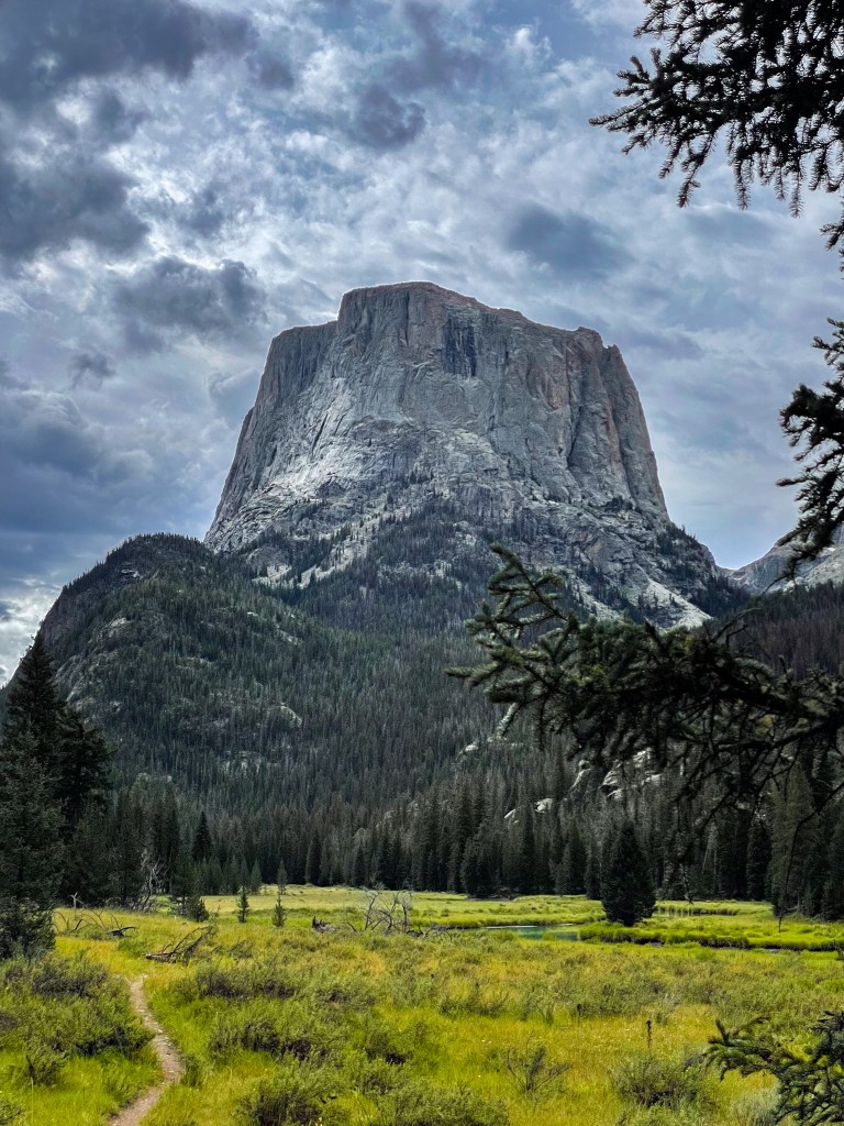 A towering rocky mountain stands prominently against a cloudy sky, surrounded by lush green meadows and forests.