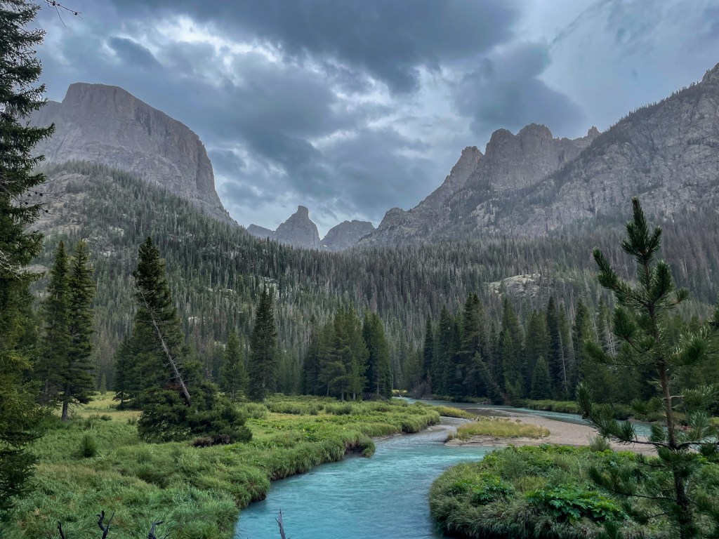 A tranquil river flowing through a lush green valley surrounded by towering mountains under a cloudy sky.