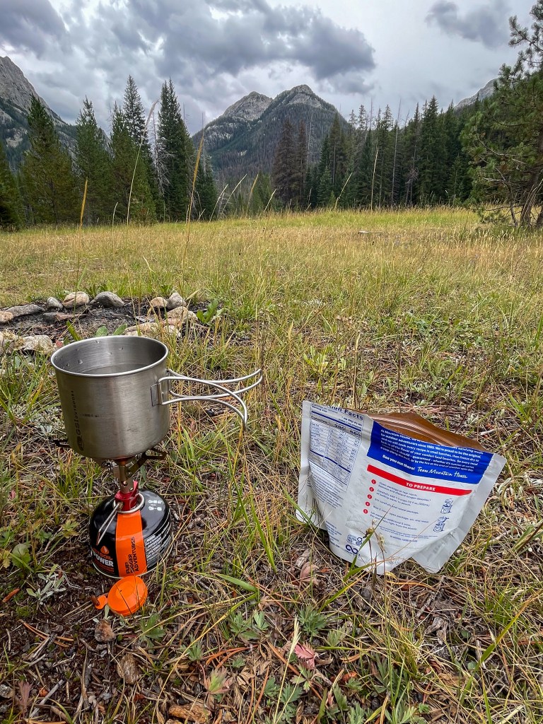 A camping stove with a metal pot is set up on grassy ground, with a food packet nearby, surrounded by trees and mountains under a cloudy sky.
