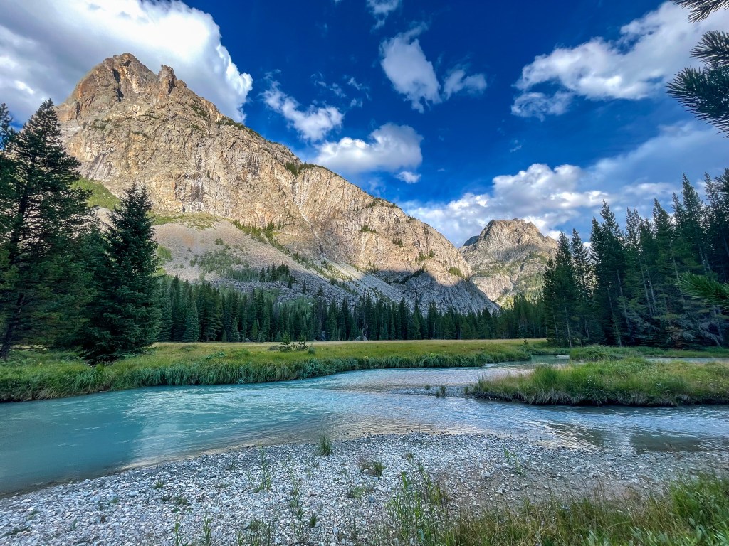A serene landscape featuring a clear river winding through a lush green meadow, surrounded by towering mountains under a partly cloudy blue sky.
